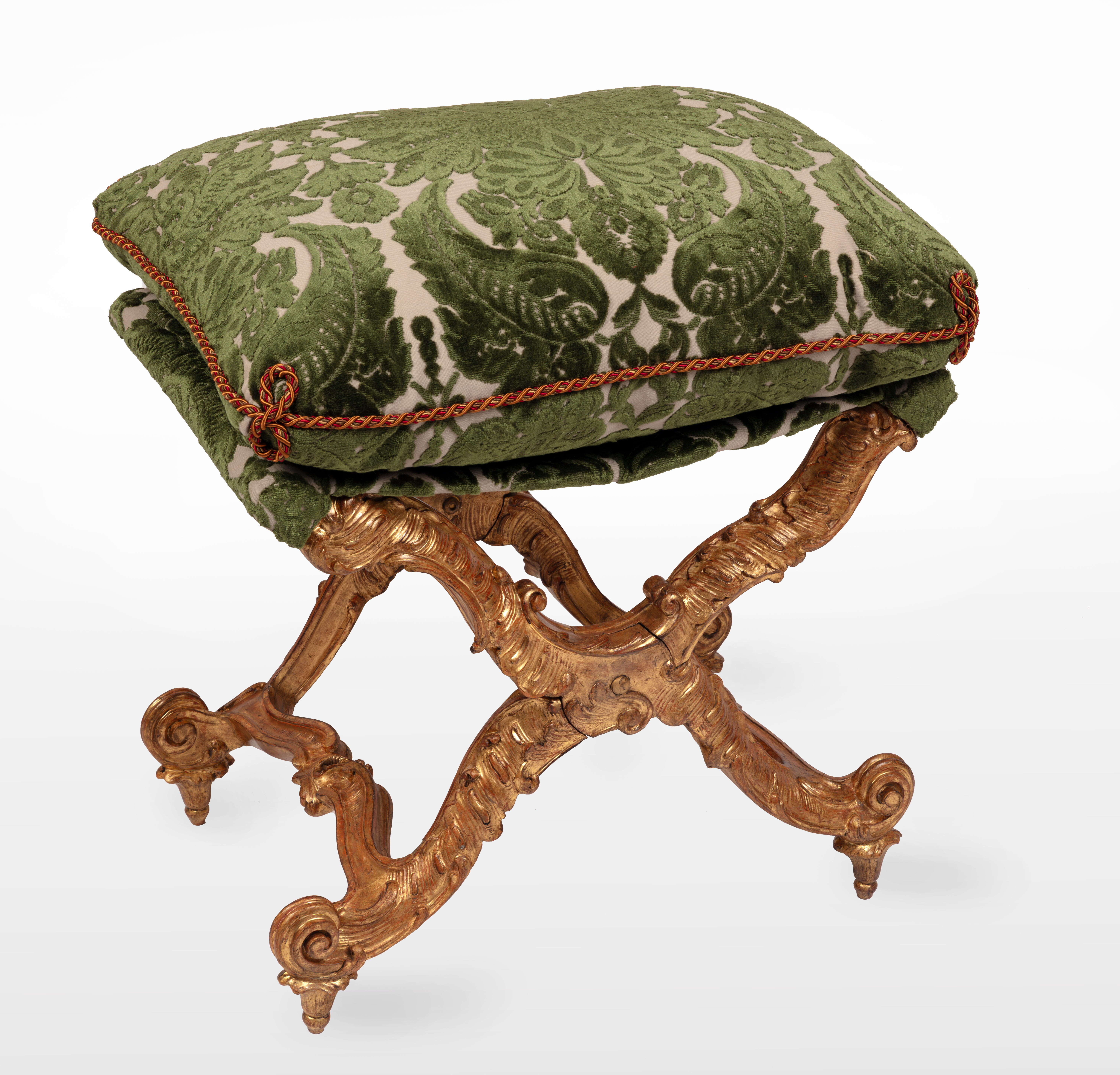 An Italian giltwood carved pliant, with a rectangular loose cushion seat in a green damask and trimming, above ‘X'-frame baluster supports, carved with scrolls, joined by shaped stretchers, on inward scrolled feet.